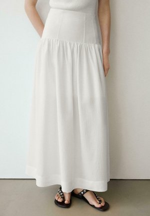 Frau trägt einen weißen, hoch taillierten, knöchellangen Rock, kombiniert mit schwarzen Sandalen mit silbernen Nieten.