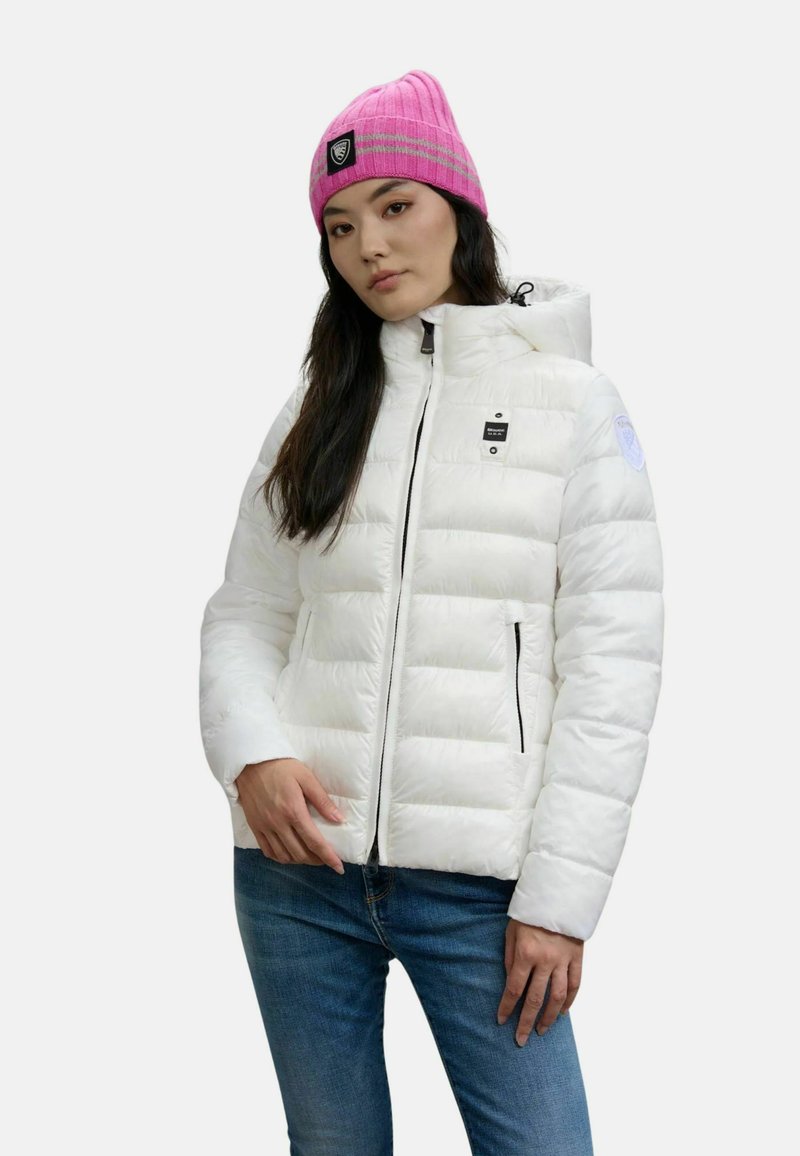Giacca puffer bianca con design a quadri orizzontali, apertura con cerniera, cappuccio e dettagli neri. Abbinata a un berretto lavorato a maglia di un vivace colore rosa.