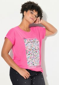 Růžové tričko s krátkým rukávem s květinovým vzorem a nápisem "ÚŽASNÉ", spárované s tmavými džínami ozdobenými perlami.