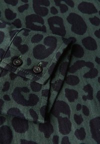 Tessuto verde con motivo a stampa leopardata scura, caratterizzato da una piega al polsino con due bottoni neri e dettagli di cucitura a contrasto.