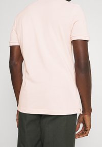 Hombre vistiendo una camiseta polo de manga corta rosa claro y pantalones oscuros, mostrado desde la espalda contra un fondo simple.