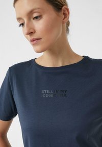 Granatowy t-shirt wykonany z miękkiego materiału, z okrągłym dekoltem i nadrukowanym tekstem, który brzmi "WCIĄŻ W MOJEJ IKONICZNEJ ERZE" w ciemnoszarym.