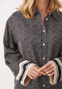 Chemise à motifs noir et blanc avec design chevrons, fermeture à boutons à l'avant et poignets rayés avec accents crème. Tissu doux, coupe décontractée.