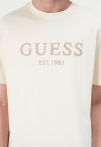 Kratka krem majica s kratkim rukavima s velikim svijetlosmeđim izvezenim logotipom "GUESS" i detaljem "EST. 1981" u središtu. Mekani pamučni materijal.