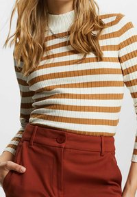 Femme portant un haut à manches longues côtelé à rayures marron et blanches, rentré dans un pantalon taille haute couleur rouille avec un bouton et des passants pour ceinture.