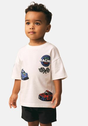 Egy kisgyermek fehér pólót visel, amelyen versenyautók, kockás zászlók és a "Kész egy kis versenyző" felirat látható, fekete rövidnadrággal párosítva.