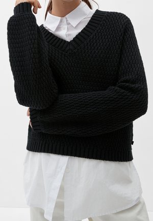 Personne portant un pull noir texturé en V par-dessus une chemise blanche boutonnée, bras croisés, sur un fond clair uni.