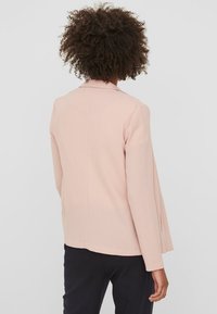Blazer rosa chiaro con colletto, vestibilità su misura e maniche lunghe. Presenta una texture liscia e un dettaglio di apertura sul retro. Adatto per abbigliamento casual o formale.