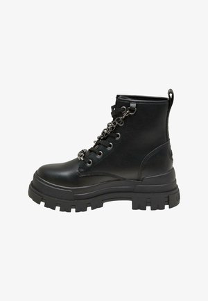 Person trägt schwarze klobige Stiefel mit Ketten, Beine ausgestreckt außerhalb einer offenen Autotür auf einem sonnigen Bürgersteig nahe einer weißen Backsteinmauer.