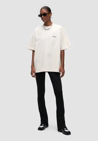 T-shirt oversize color crema con maniche corte e dettaglio del logo, abbinato a pantaloni neri a zampa d'elefante e scarpe nere. Gli accessori includono occhiali da sole e collana.