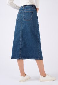 Denim-Midirock in Dunkelblau mit gerader Silhouette, ausgestattet mit zwei Gesäßtaschen und einem Saum knapp über den Knöcheln. Kombiniert mit weißen Sneakers.