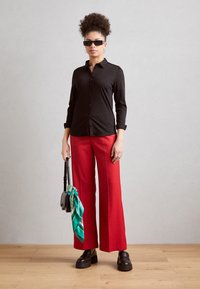 Camicia nera a bottoni, pantaloni rossi a gamba larga, scarpe nere spesse, borsa nera sottile con un accento di sciarpa turchese, e occhiali da sole quadrati. Texture lisce.