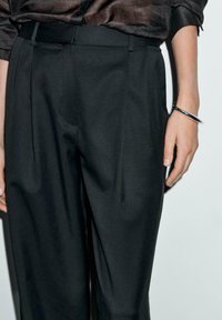 Black tailored trousers with a smooth texture, featuring a pleated front, belt loops, and a side pocket. Hand wearing a silver bracelet.