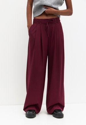 Personne portant un pantalon bordeaux taille haute à jambes larges avec des plis avant, des chaussures noires épaisses, et un top gris court sans manches.