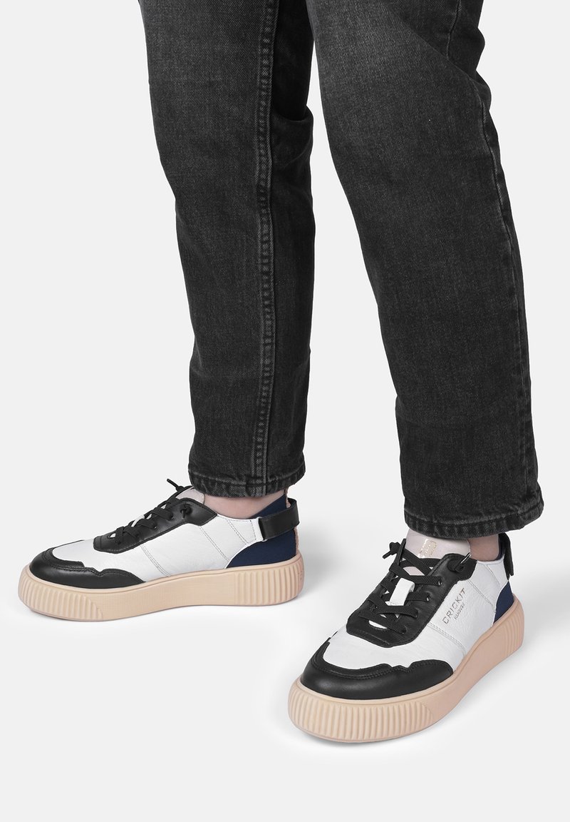 Baskets noires et blanches avec une semelle épaisse beige, un tissu texturé, des lacets noirs et un accent bleu sur le talon. Portées avec un jean sombre.