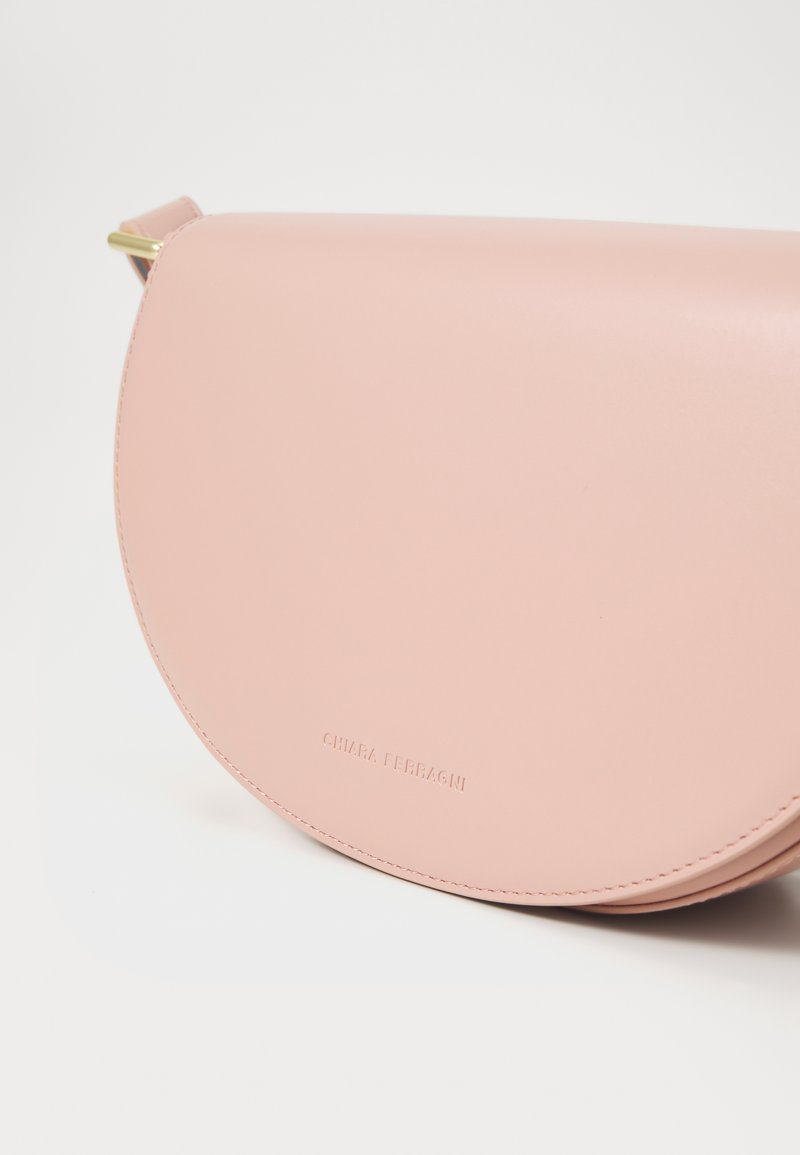 Rózsaszín bőr keresztpántos táska ívelt fedelemmel, diszkrét varrással és dombornyomott logóval. Aranyszínű díszítéssel rendelkezik a pánton.