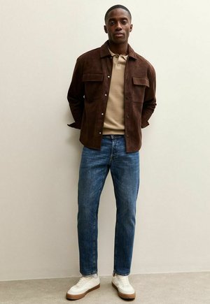 Giacca in camoscio marrone con due tasche sul petto, polo beige, jeans blu e sneakers bianche con suola marrone su sfondo chiaro.