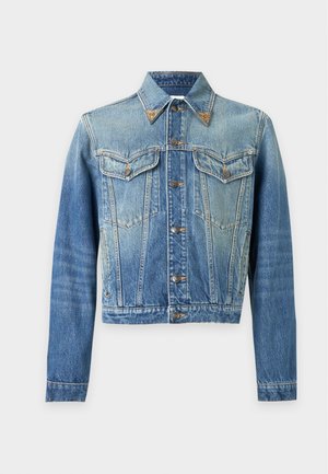 Junger Mann trägt eine blaue Jeansjacke, buntes Hemd, schwarze Hose mit Lederdetail und schwarze Schuhe mit goldenen Akzenten, steht vor weißem Hintergrund.