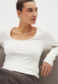 Top bianco a coste con maniche lunghe e scollatura a barca, indossato con pantaloni scuri. Il modello ha orecchini a cerchio argentati e un anello, seduto su una superficie grigia.