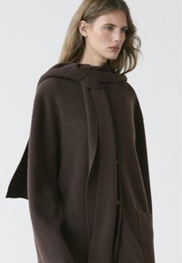 Femme aux longs cheveux châtain clair portant un manteau en laine brun foncé surdimensionné avec de grandes poches avant et une écharpe assortie nouée autour du cou.