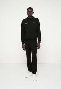 Czarna bluza z kapturem z przednią kieszenią i detalem logo, w połączeniu z czarnymi dresami. Model nosi białe sneakersy. Gładki materiał, luźny krój.