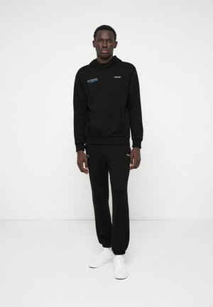 Čierna mikina s kapucňou a predným vreckom, s logom, skombinovaná s čiernymi joggermi. Model má na sebe biele tenisky. Hladký materiál, voľný strih.