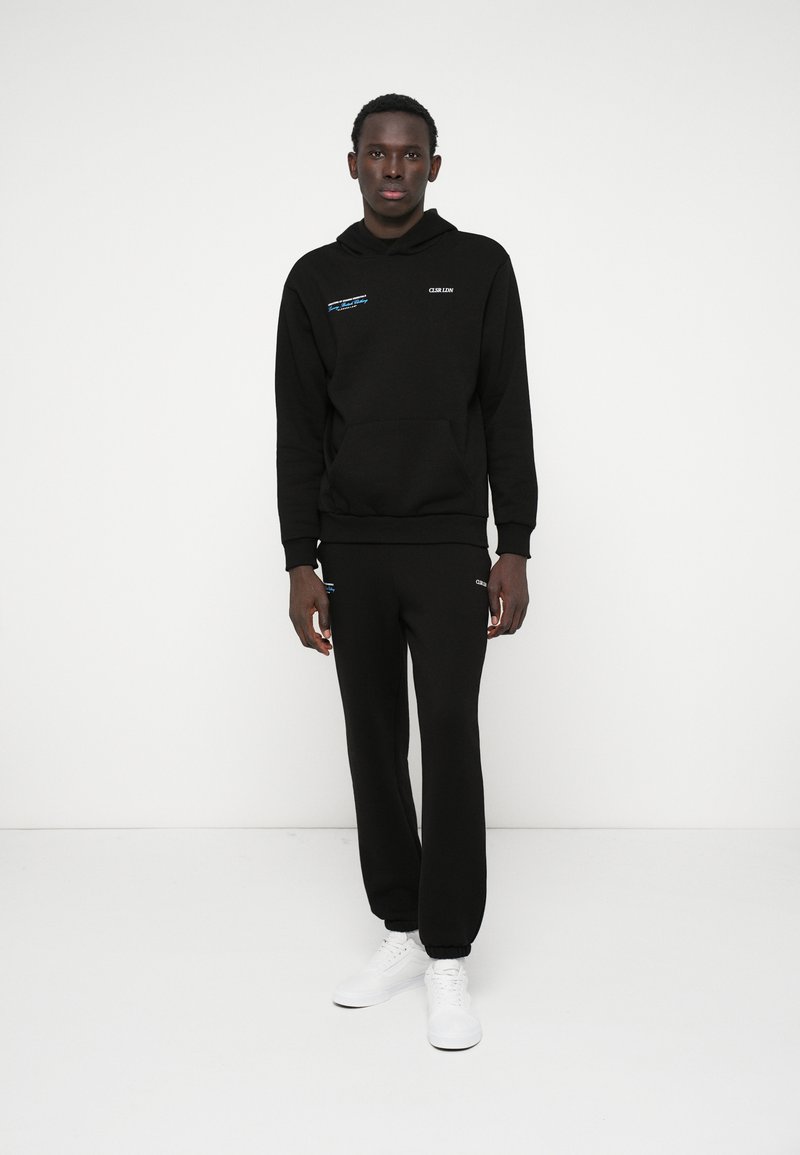 Czarna bluza z kapturem z przednią kieszenią i detalem logo, w połączeniu z czarnymi dresami. Model nosi białe sneakersy. Gładki materiał, luźny krój.