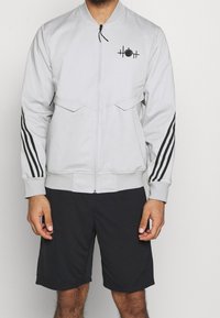Man wearing light gray zip-up jacket with black stripes on sleeves and black shorts, standing against a plain background.