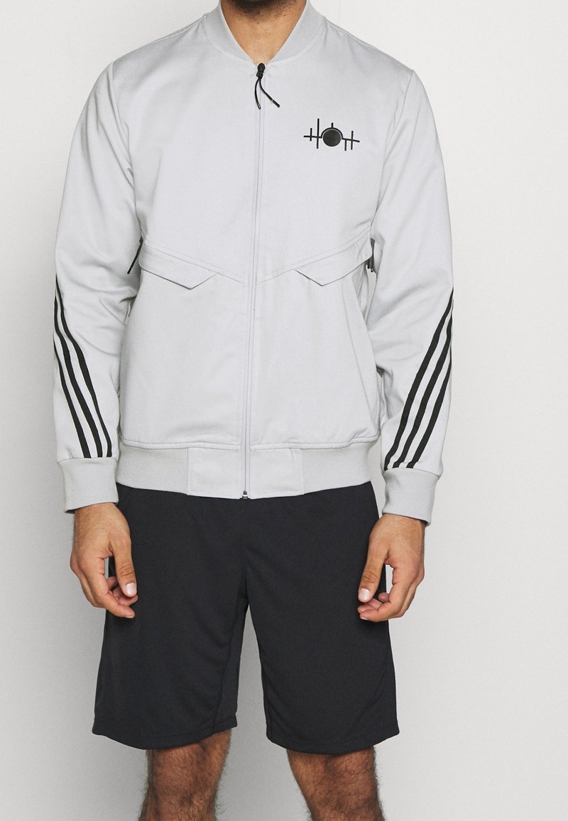 Man wearing light gray zip-up jacket with black stripes on sleeves and black shorts, standing against a plain background.