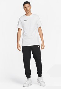 Weißes Baumwoll-T-Shirt mit einem kleinen schwarzen Nike-Logo, kombiniert mit schwarzen Jogginghosen und weißen Sportschuhen. Einfaches Design und lässige Passform.