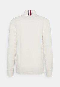 Maglione bianco a maglia con colletto alto. Presenta una striscia rossa, bianca e nera al collo e polsini e orlo a costine. Vista dal retro.