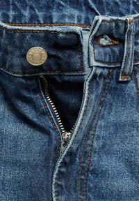 Denim kavbojke z vidno kovinsko gumbom in zadrgo. Imajo oranžno šivanje in temno modro pranje. Prikaz blizu pasu in območja zadrge.