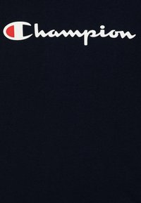 Marineblå t-shirt i bomull. Har en vit och röd "Champion"-logotyp tryckt över bröstet i en böjd och kursiv stil.
