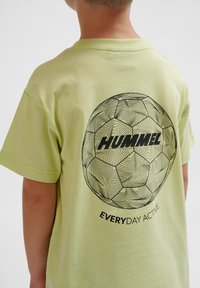 Lys grøn bomulds-t-shirt med et sort grafisk design af en fodbold og teksten "HUMMEL EVERYDAY ACTIVE" på bagsiden. Korte ærmer, rund hals.