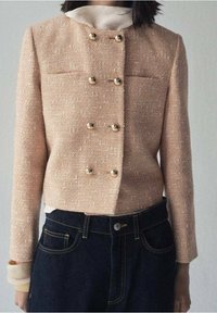 Blazer rose texturé avec un col montant, un design croisé et des boutons dorés, associé à un jean en denim foncé.