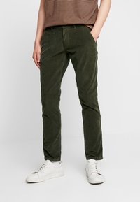 Zielone spodnie sztruksowe o wąskim kroju, z kieszeniami z przodu, zestawione z białymi trampkami i brązową bluzką w paski.