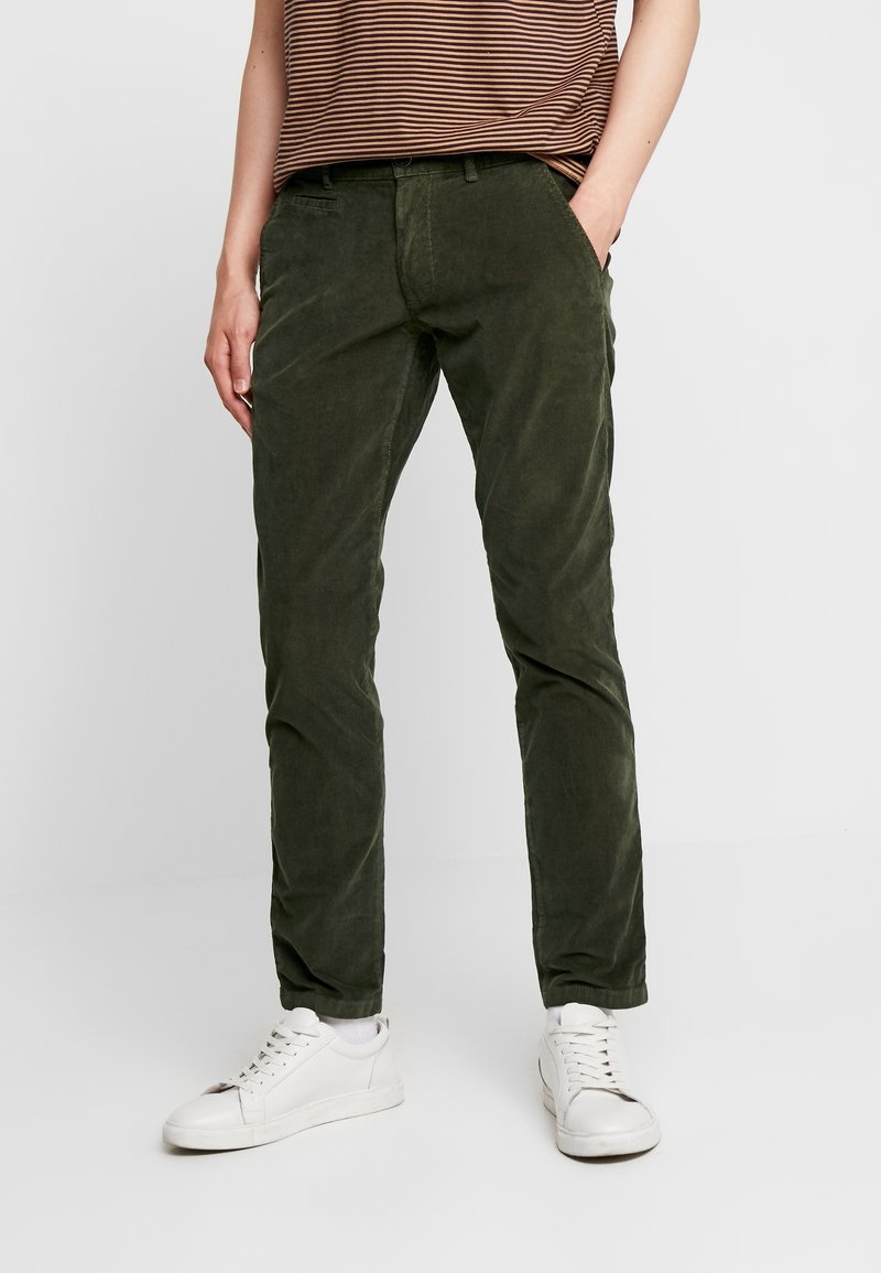 Zielone spodnie sztruksowe o wąskim kroju, z kieszeniami z przodu, zestawione z białymi trampkami i brązową bluzką w paski.