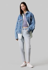 Chaqueta de denim azul grande sobre una camisa verde clara, combinada con un top corto de canalé morado, jeans claros y zapatillas blancas.