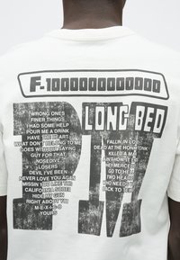 T-shirt branca de algodão com texto gráfico preto em negrito nas costas, apresentando títulos de canções e letras em vários tamanhos de fonte. Textura macia.