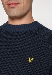 Maglione lavorato a maglia di un intenso blu navy con un motivo testurizzato, presenta un collo rotondo a coste e un piccolo logo di un uccellino giallo sul lato sinistro del petto.