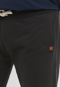 Sorte joggebukser laget av mykt stoff, med ribbestrikket midjebånd, snor i midjen, sidelommer og en liten oransje logo-lapp.