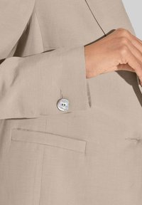 Lichtbeige blazer met een gestructureerde afwerking. Heeft een knoop van parelmoer met twee kleine gaatjes en een zijzakdetail.