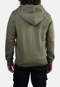 Homme portant un sweat à capuche vert olive et un pantalon noir, se tenant de dos face à l'appareil photo sur un fond uni.