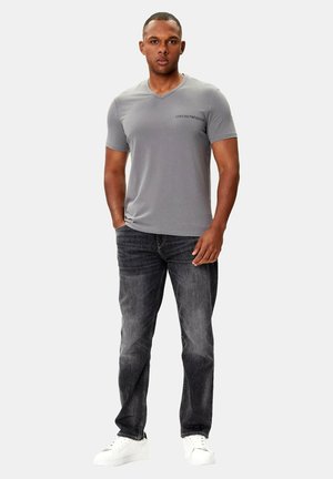 T-shirt grigia a scollo a V con maniche corte, realizzata in tessuto liscio, abbinata a jeans denim scuro e sneakers bianche. Logo evidente sul petto.