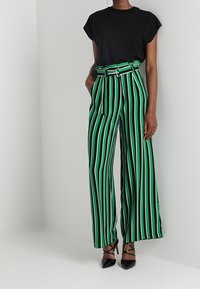 Svart kortärmad topp kombinerad med högmidjade grön- och svartrandiga vida byxor, med ett knytbälte och blanka svarta klackar.