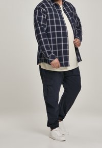 Marinblå rutigt knäppt skjorta över en ljus krämfärgad t-shirt, parat med marinblå cargobyxor och vita sneakers.