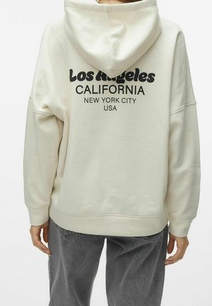 Persona che indossa una felpa color crema con la scritta "Los Angeles California New York City USA" sulla schiena, abbinata a jeans grigi.