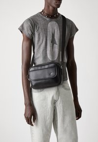 Borsa a tracolla rettangolare nera realizzata in materiale liscio, con una tasca frontale con zip, tracolla regolabile e un piccolo logo.