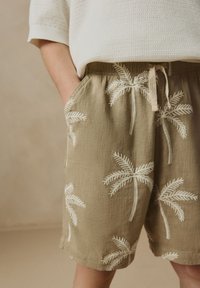 Beige korte broek met witte palmboomborduurwerk, gemaakt van lichtgewicht stof. Voorzien van een elastische tailleband en zijzakken.