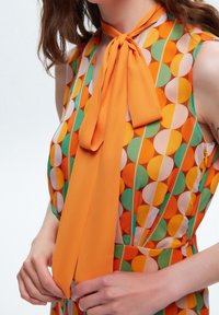 Vestido sin mangas con patrones circulares coloridos en naranja, verde y amarillo, y un prominente detalle de lazo naranja en el escote.